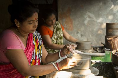 Two women shaping clay on potter's wheels, one trimming a pot with a metal tool