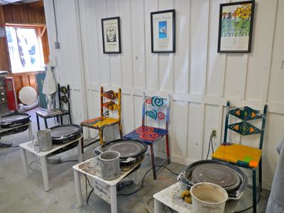 A row of electric potter's wheels with water buckets in front of hand-painted wooden chairs, in a white-paneled studio with framed posters